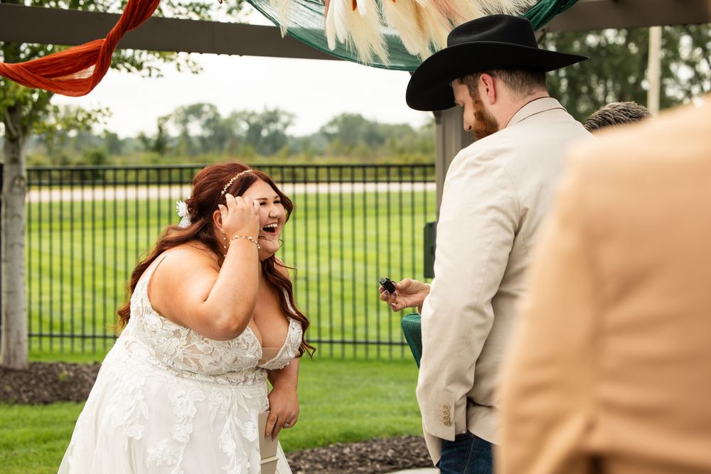 Joyful bride in a white lace gown wipes happy tears during an outdoor wedding ceremony as guests in cowboy hats look on.