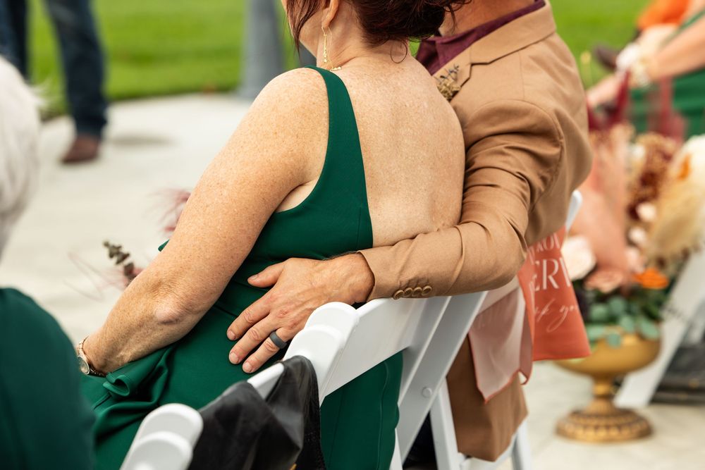 Woman in an emerald green dress seated on a white chair while a person in a tan jacket wraps an arm around her at an outdoor wedding.