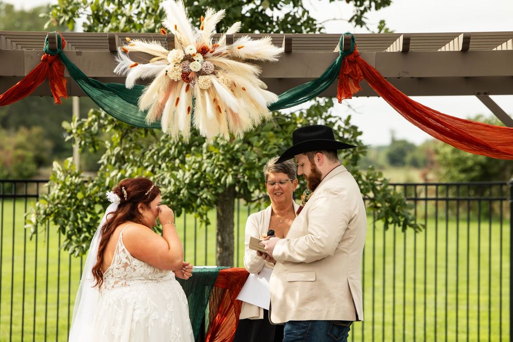 Bride in a white lace gown wipes her tears as an officiant and a bearded groom in a black hat stand under a boho pergola with pampas decor.