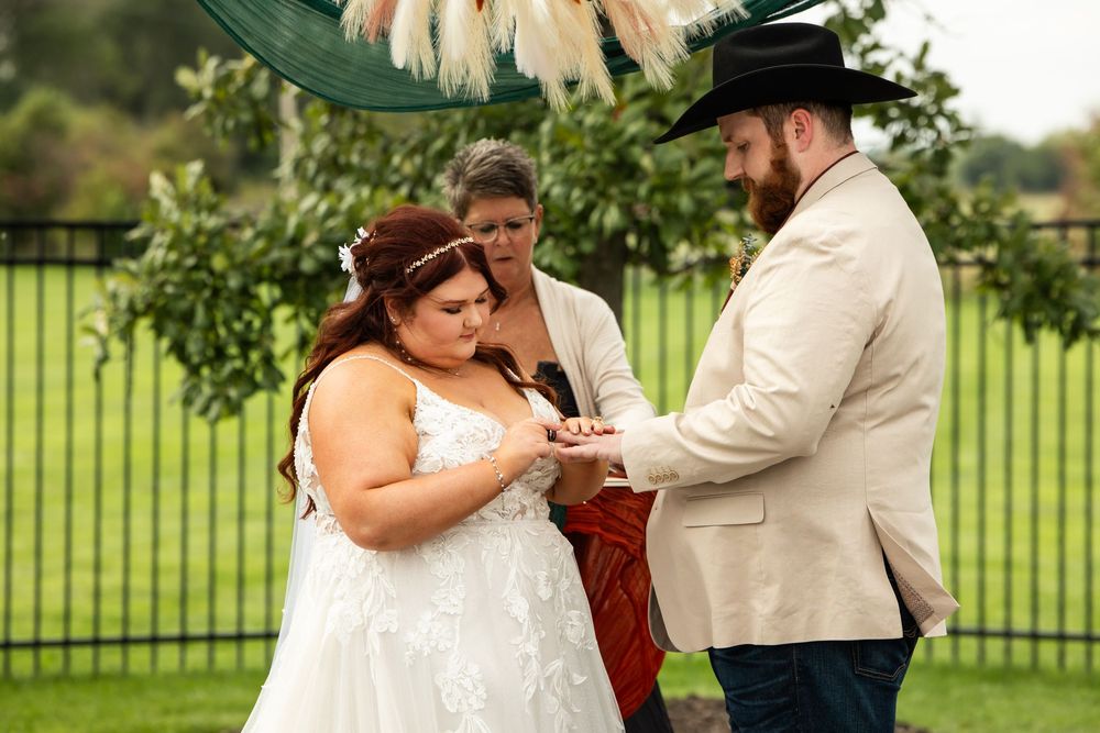 Bride in a white lace gown and groom in a cowboy hat and tan blazer exchange rings during an outdoor wedding ceremony with an officiant.
