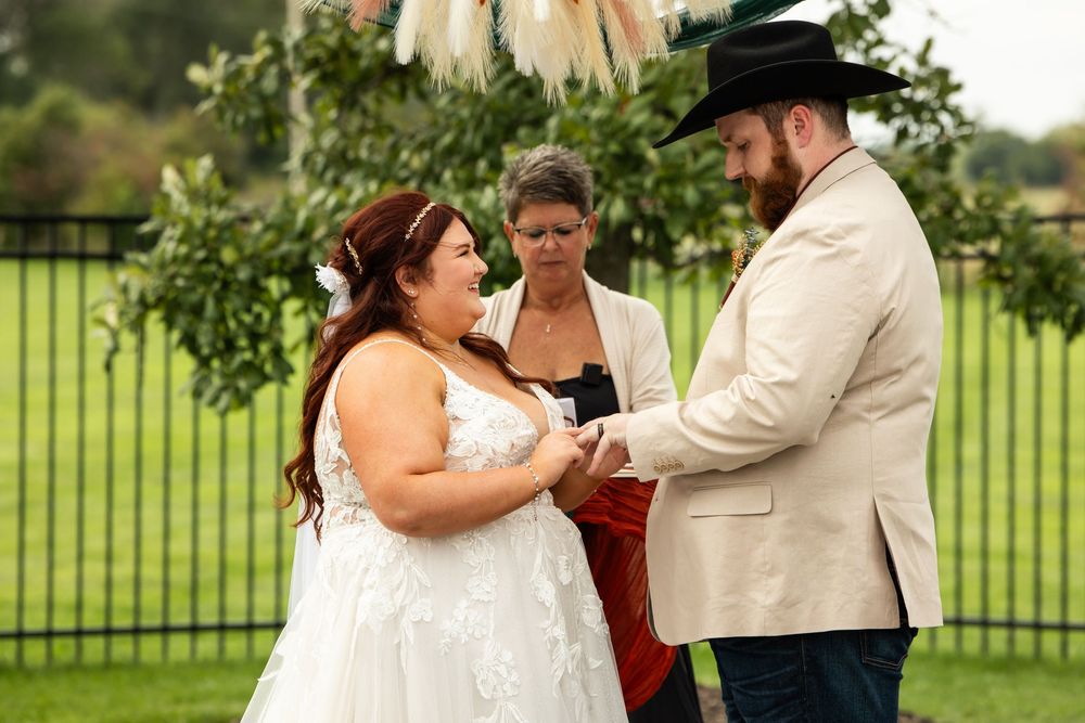 A bride in a lace wedding dress and a bearded groom in a cowboy hat hold hands during their outdoor vow exchange beside a smiling officiant.