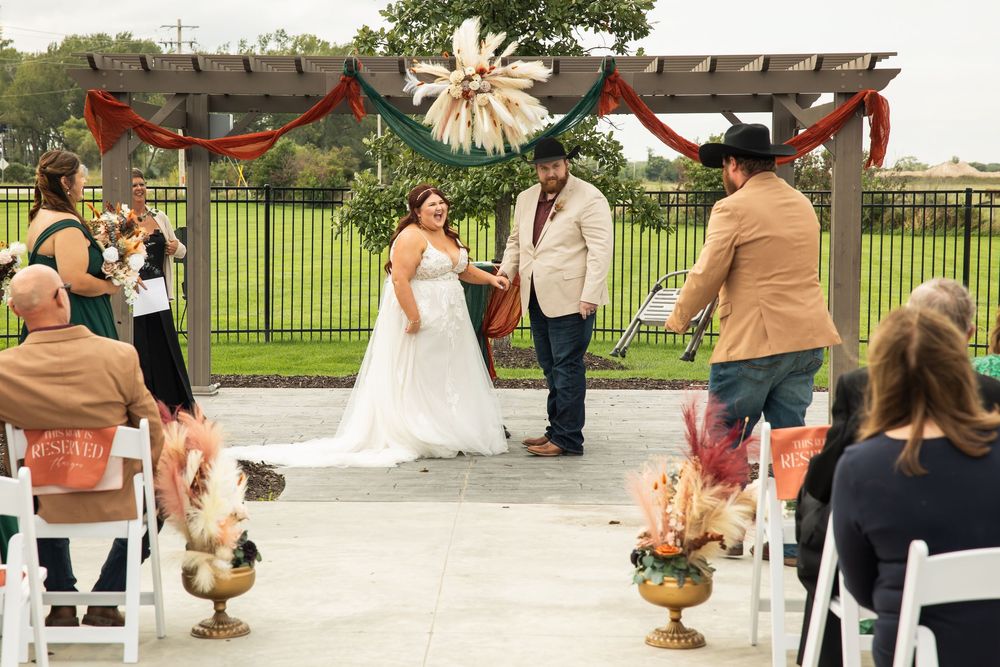 Bride in a white gown and groom in a vest stand under a pergola with pampas and rust draping during an outdoor boho wedding ceremony.