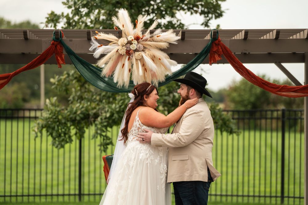 Bride in a white lace gown embraces a groom in a tan suit and black cowboy hat beneath a boho pampas floral arch with rust and teal drapes.