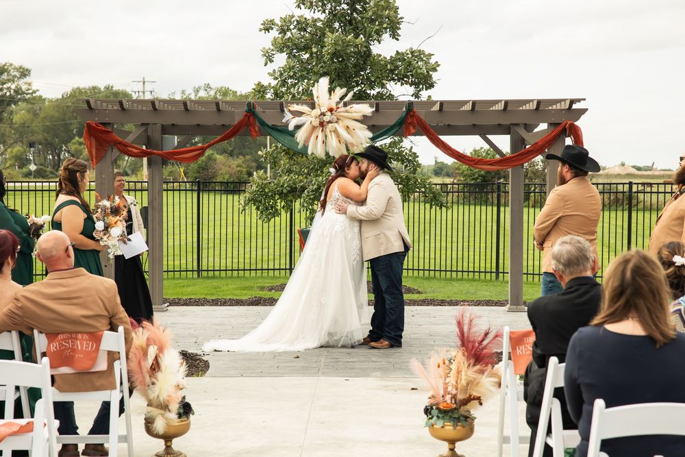 Bride in a white gown and groom in a cowboy hat embrace under a rustic wooden pergola at an outdoor wedding ceremony with guests watching.