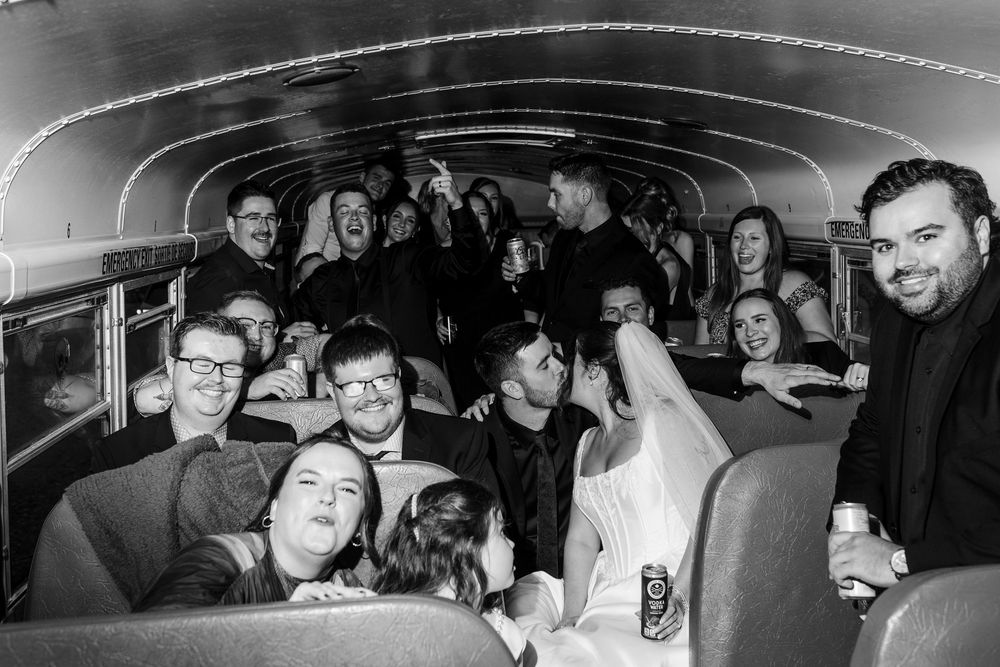 Wedding party in formal attire laughing and posing together inside a crowded school bus, with a bride in a white gown in the foreground.