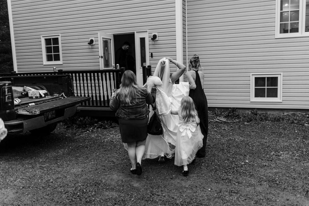 Black and white photo of bridesmaids helping a bride in a long white gown walk toward a house, with a pickup truck parked nearby.