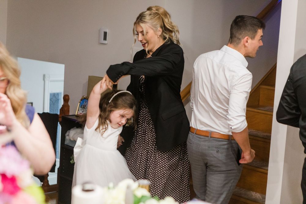 A smiling woman in a black blazer dances with a little girl in a white dress at an indoor family celebration while a man stands nearby.