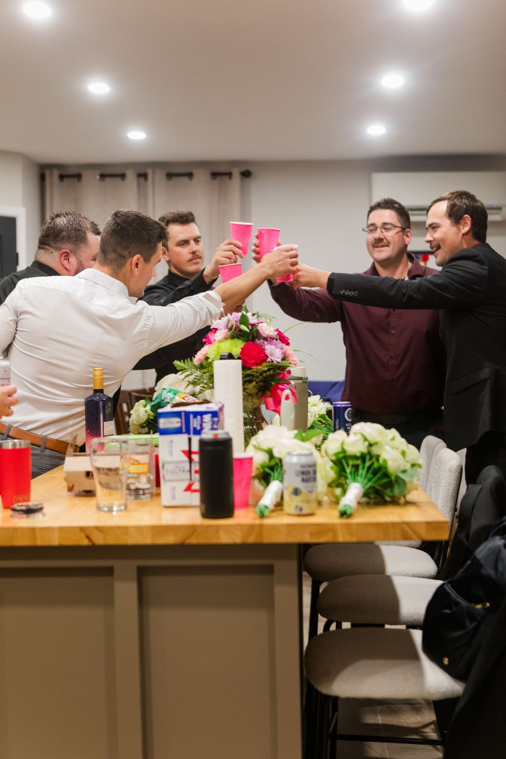 Group of men raising pink cups in a toast around a kitchen island with flower bouquets, bottles, and bar stools at a celebration.