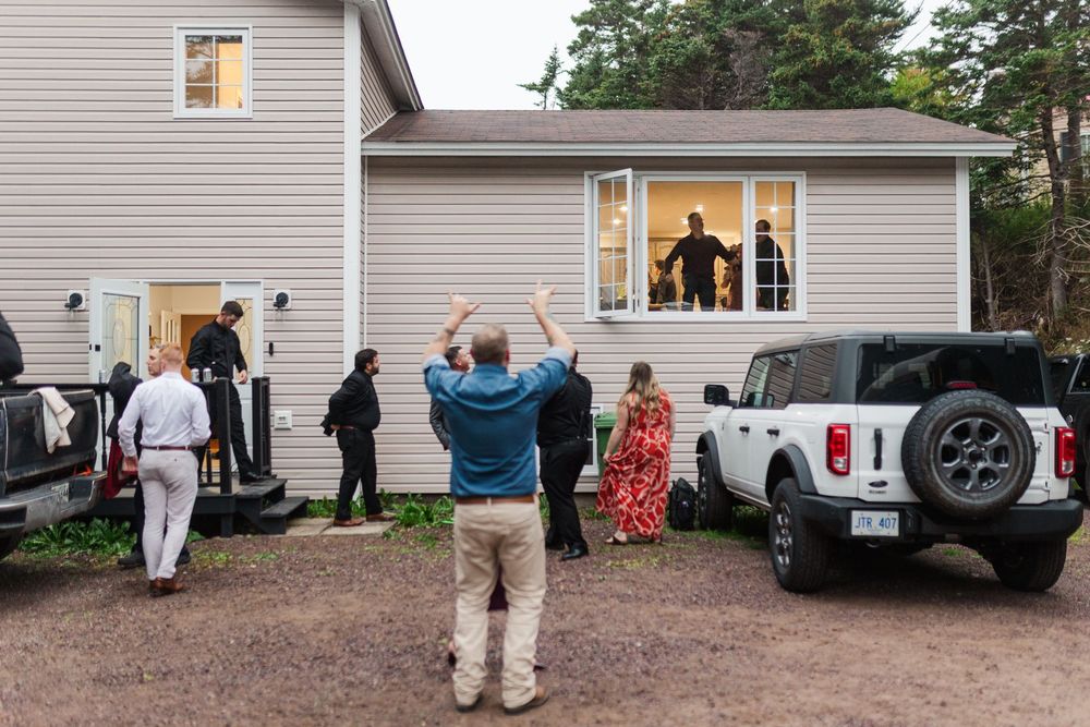 Wedding guests gather outside a beige two-story house as a man raises his arms toward people on the porch, with a white SUV parked nearby.