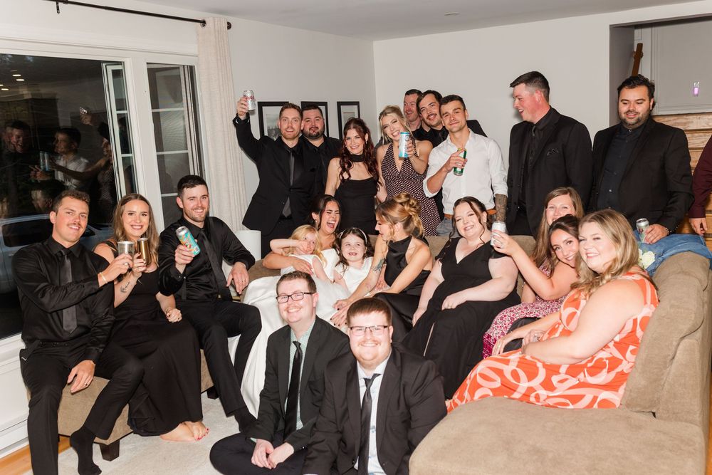 Large group of smiling friends in formal black attire posing together on couches in a bright living room at a festive party gathering.