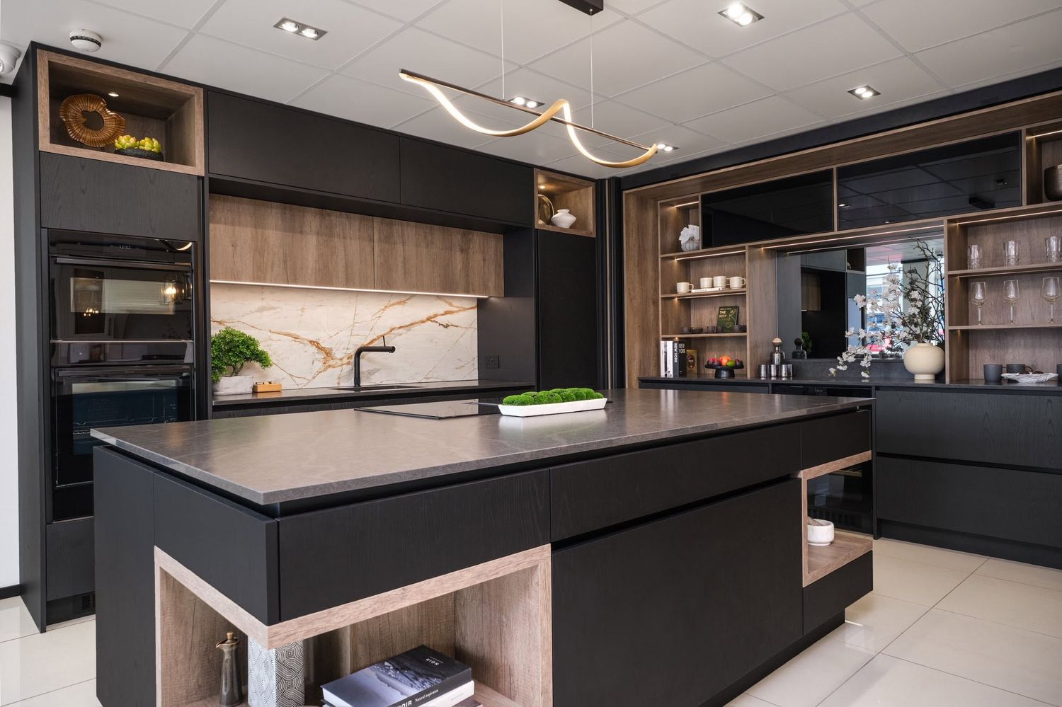 Sleek modern kitchen showroom featuring matte black cabinetry, wood accents, large island, and pendant lighting.