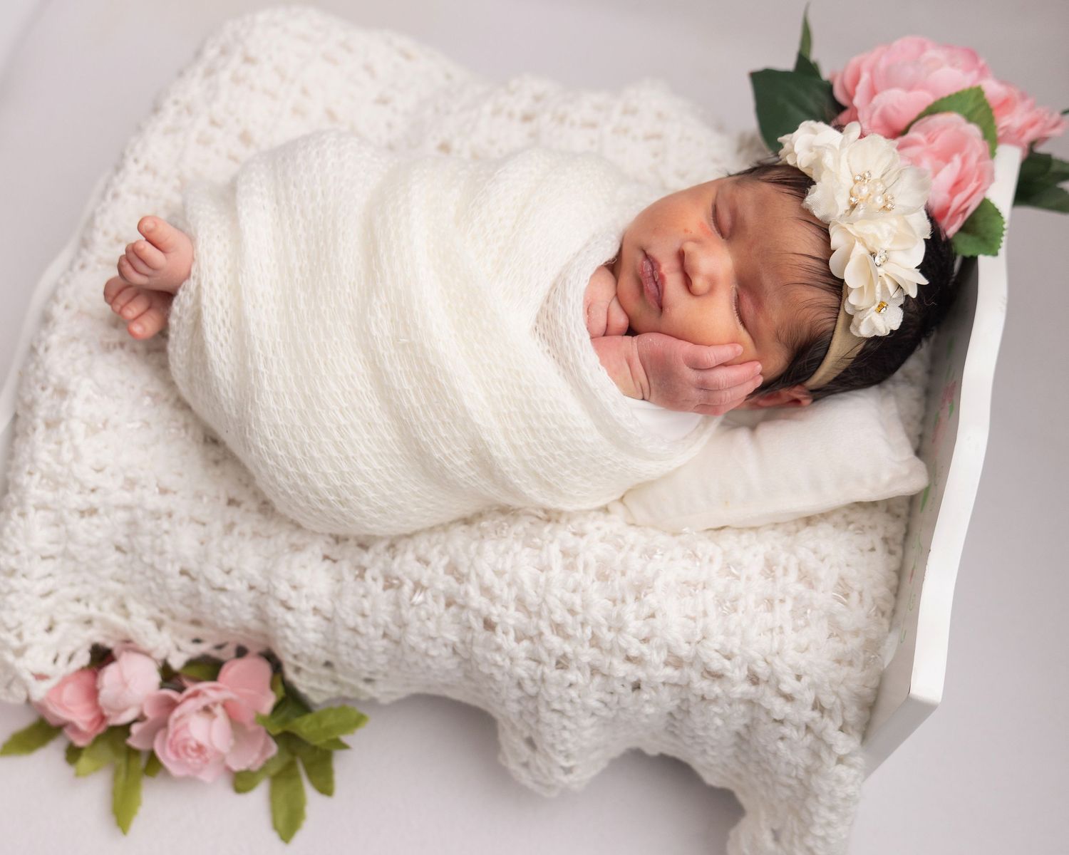 Sleeping newborn baby swaddled in white knit wrap with floral headband on crochet blanket surrounded by pink roses.