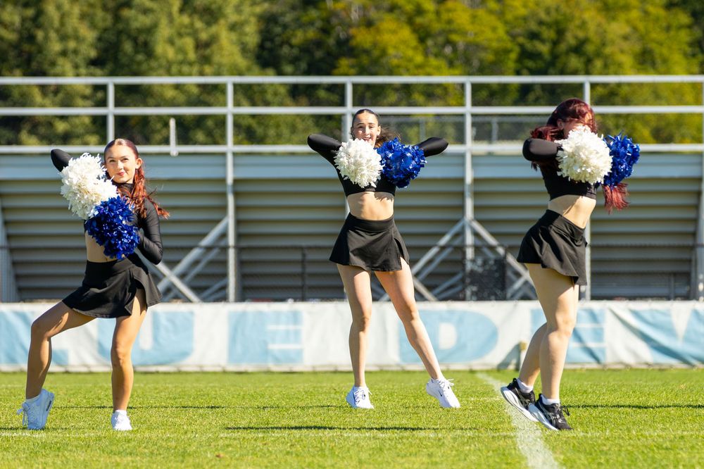 Three cheerleaders in black uniforms perform synchronized routine on football field holding blue and white pom-poms.