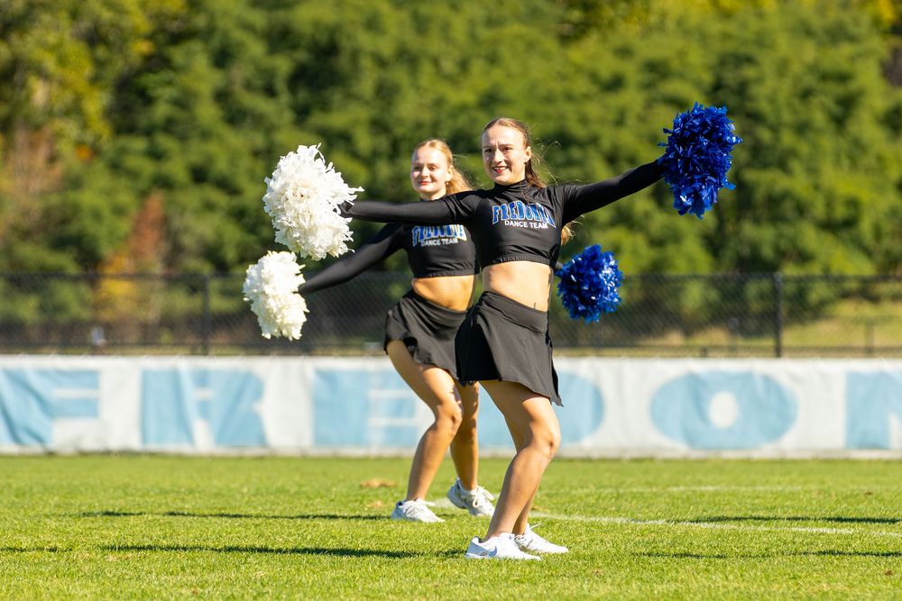 Two cheerleaders in black uniforms perform on a green field, holding blue and white pom-poms during a game.