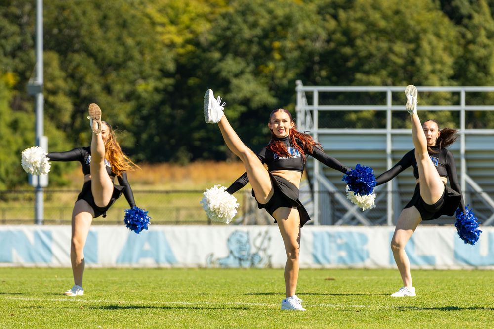 Three cheerleaders perform high kicks on a green field, holding blue and white pom-poms in matching black uniforms.