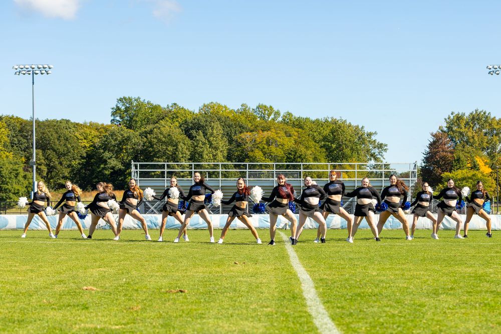 Cheerleaders performing synchronized routine on green football field under blue sky with trees in background.