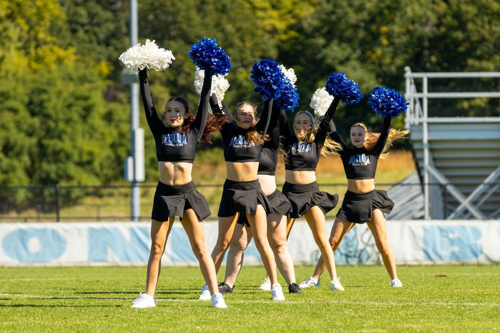 Cheerleaders in black uniforms perform with blue and white pom-poms on a green football field outdoors.