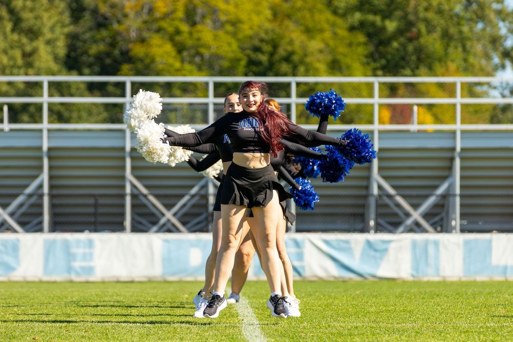 Cheerleaders perform on a green field, holding blue and white pom-poms during an outdoor sports event.