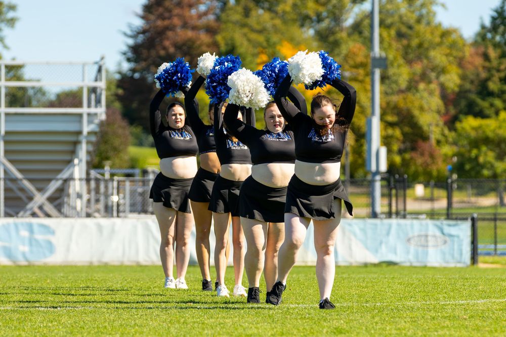 Cheerleaders in black uniforms wave blue, gold, and white pom-poms while walking across a green sports field.