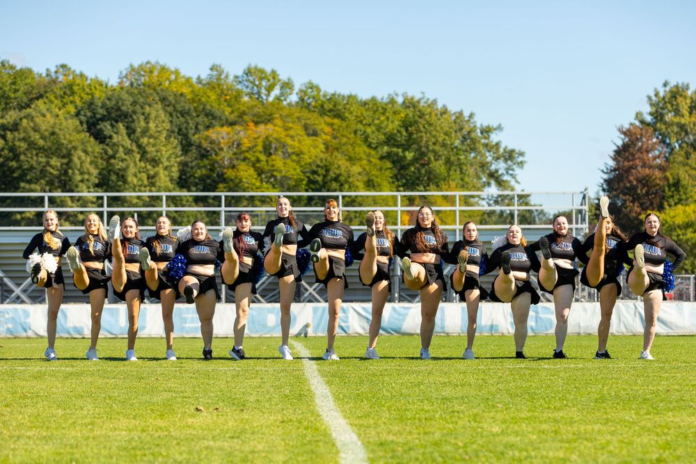 A cheerleading squad performs a synchronized high kick routine on a green football field on a sunny day.