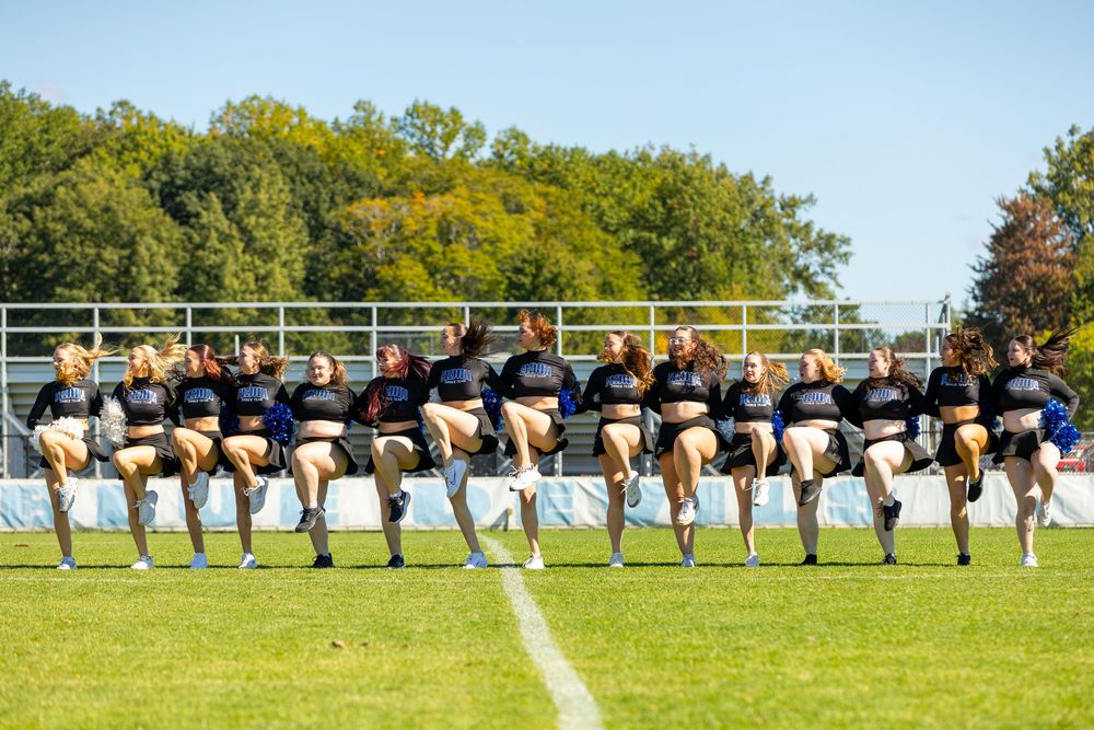 A cheerleading squad performs a synchronized kick line routine on a sunny green football field outdoors.