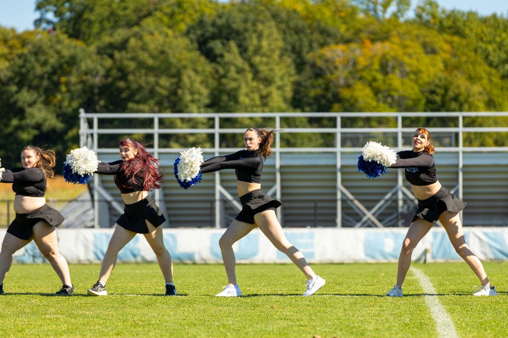 Four cheerleaders perform a dynamic routine with blue and white pom-poms on a sunny football field.