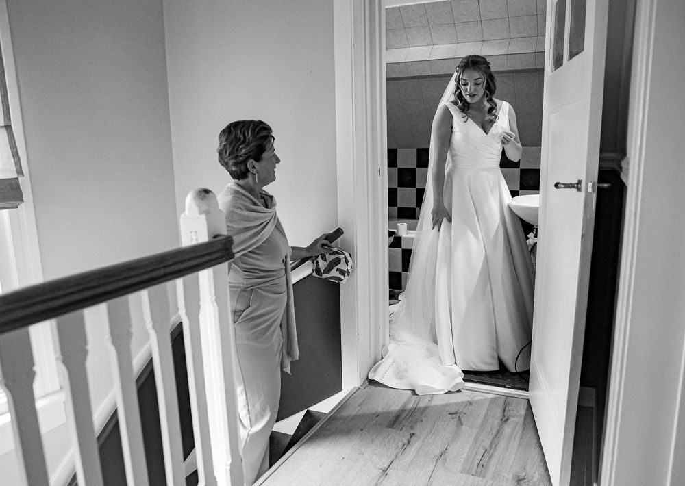 Black and white photo of a bride in a white gown standing in a doorway as a woman admires her on a staircase.