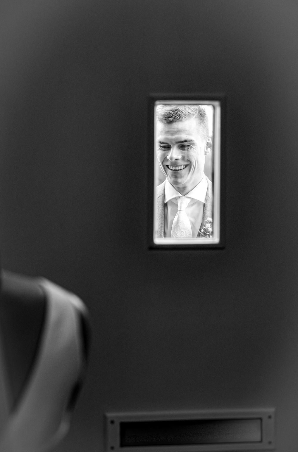 Black and white photo of a smiling groom seen through a small window, wearing a suit with a boutonniere.