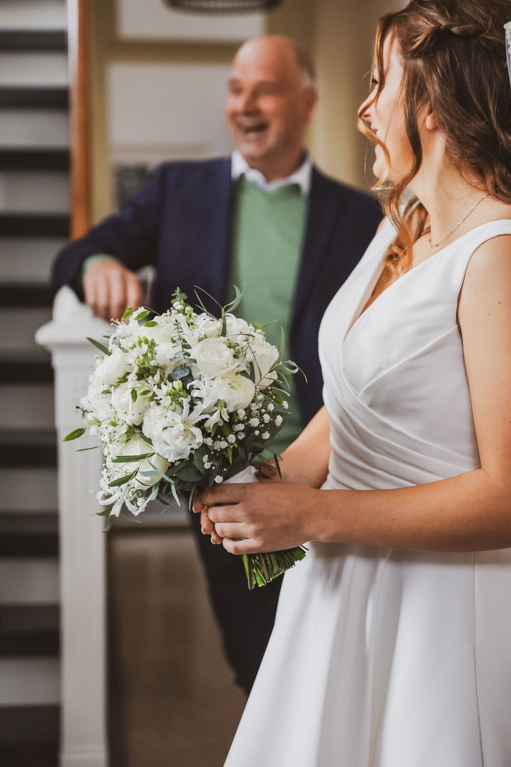 Bride in white dress holding elegant white floral bouquet with groom smiling in background at wedding venue.