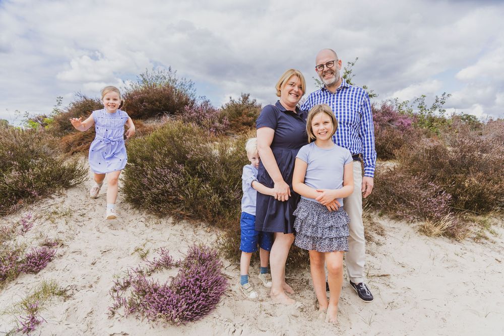 Family of five posing on sandy dunes with purple heather, smiling outdoors under a cloudy sky.