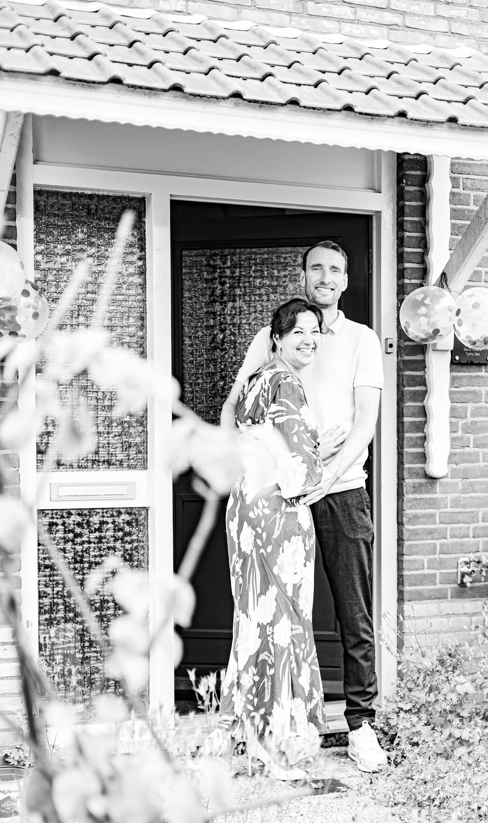Black and white photo of a smiling couple standing together in a brick home doorway, surrounded by garden plants.