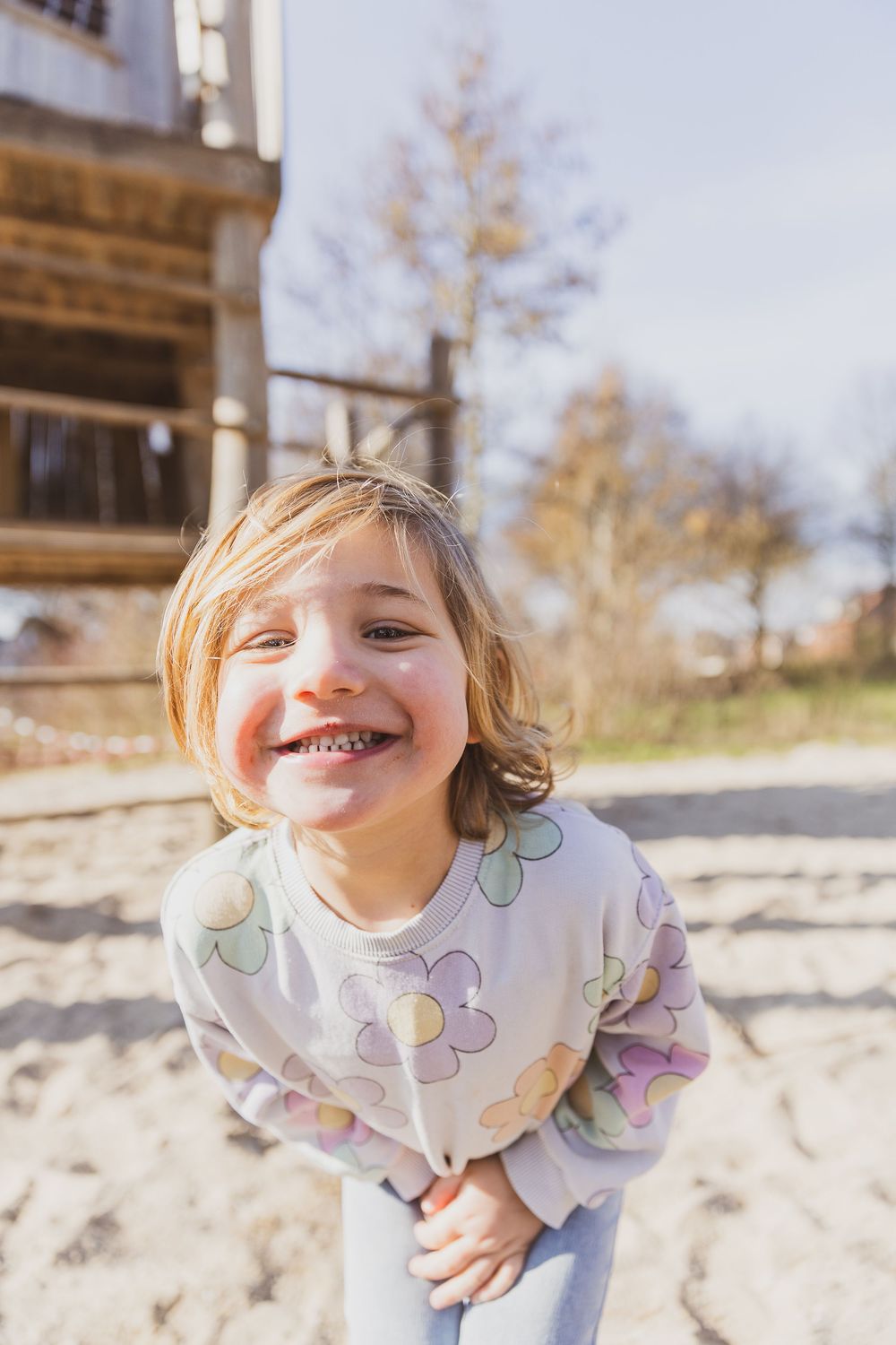 Happy young girl with blonde hair smiling outdoors, wearing a floral sweatshirt on a sunny day near a wooden building.