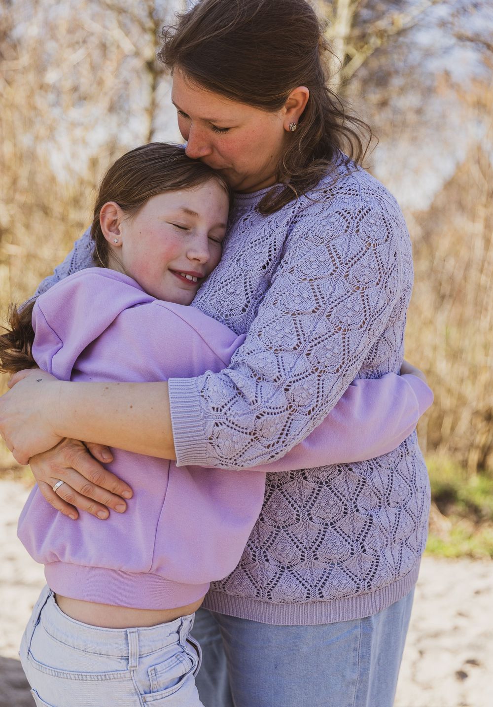 A mother and daughter share a warm embrace outdoors, both wearing matching lavender outfits in a sunny natural setting.