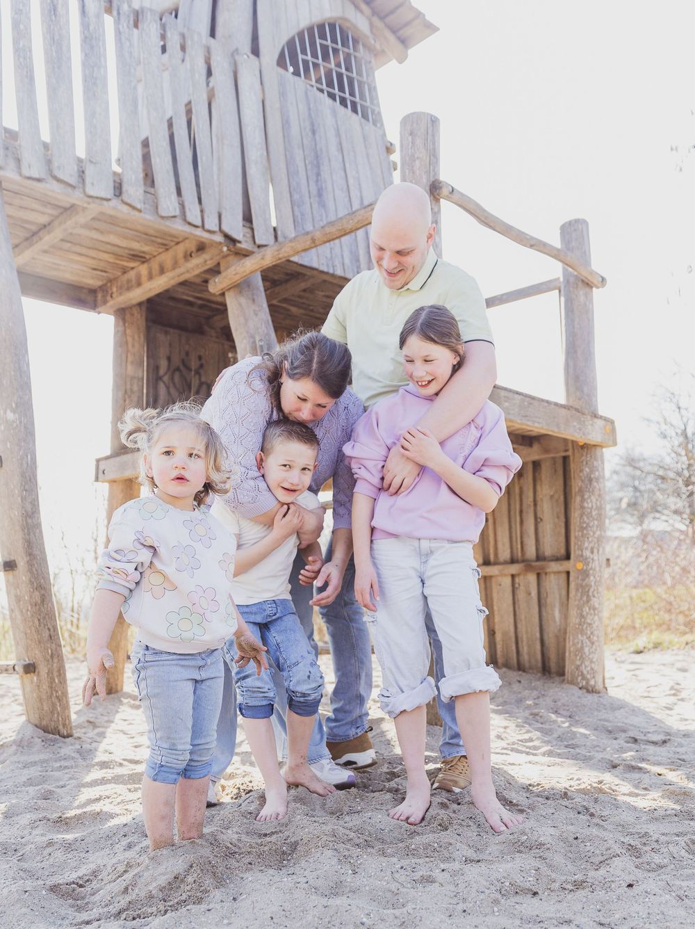 A happy family with three young children poses barefoot on sandy ground near a wooden playhouse structure.