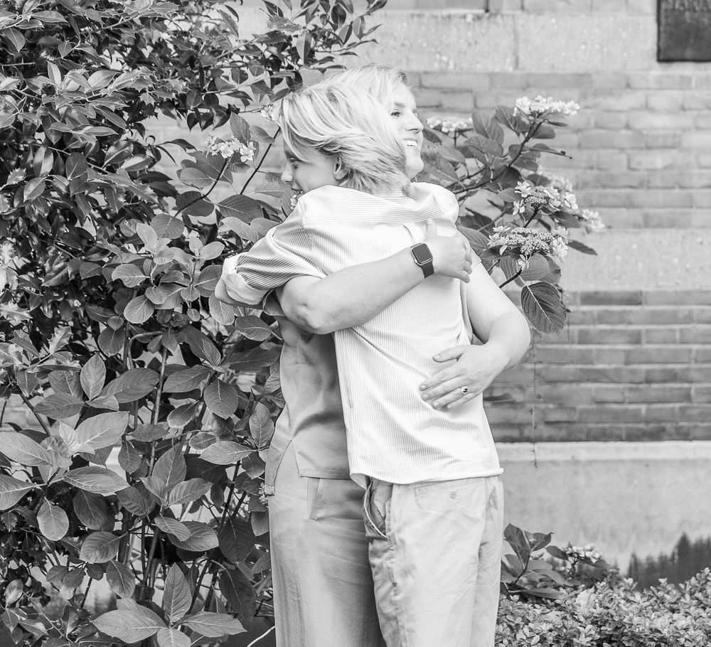 Two people share a warm embrace outdoors near lush green bushes in this black and white photo.