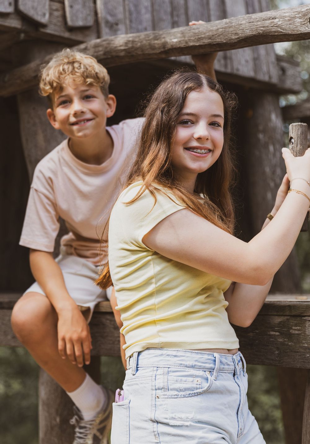 Two smiling kids playing on a wooden structure outdoors on a sunny summer day.