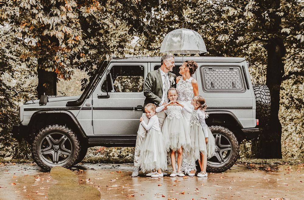 Family in formal attire poses before a gray Mercedes G-Wagon SUV outdoors in autumn.