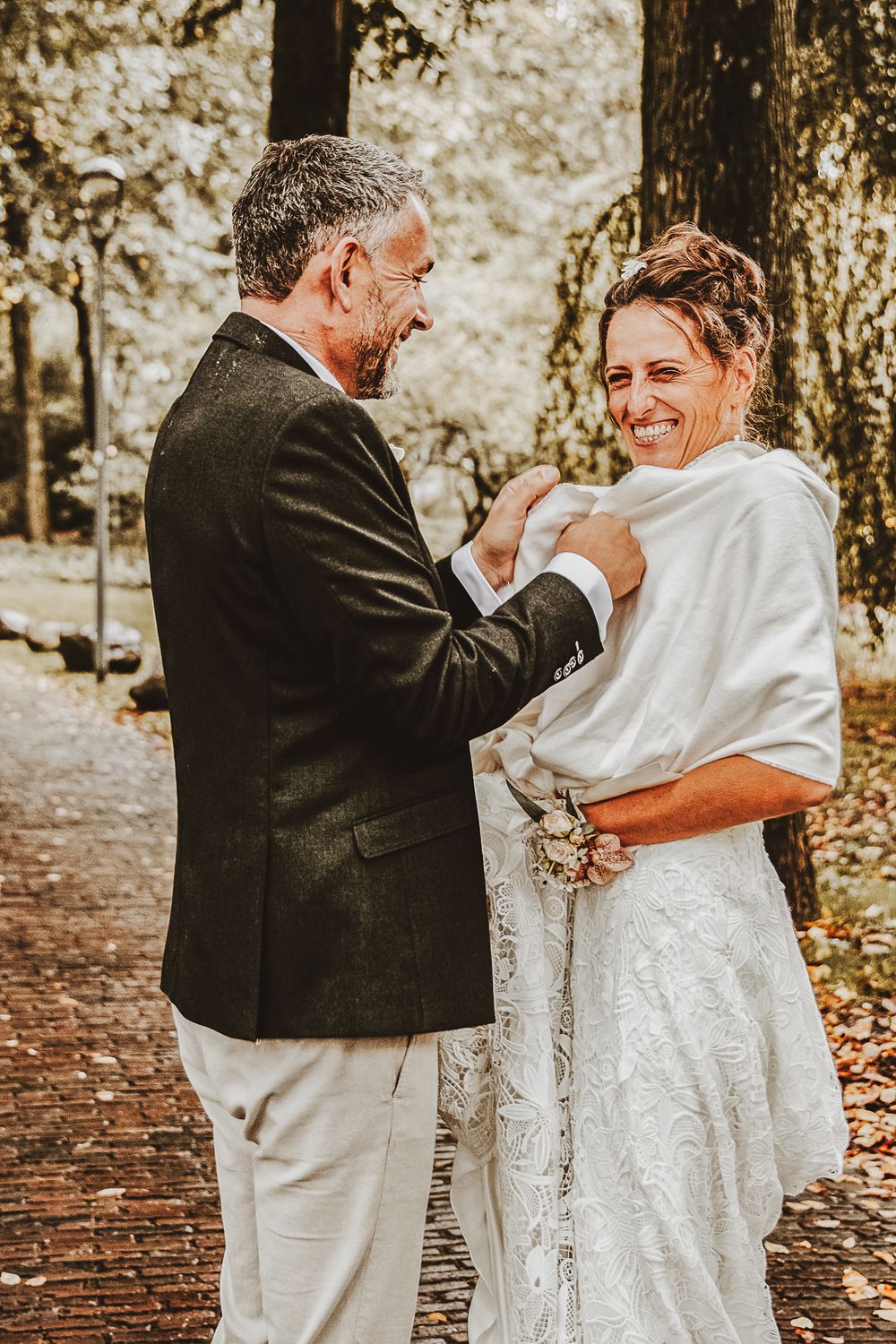 A groom adjusts a white bridal shawl on a smiling bride outdoors on a brick path surrounded by autumn trees.