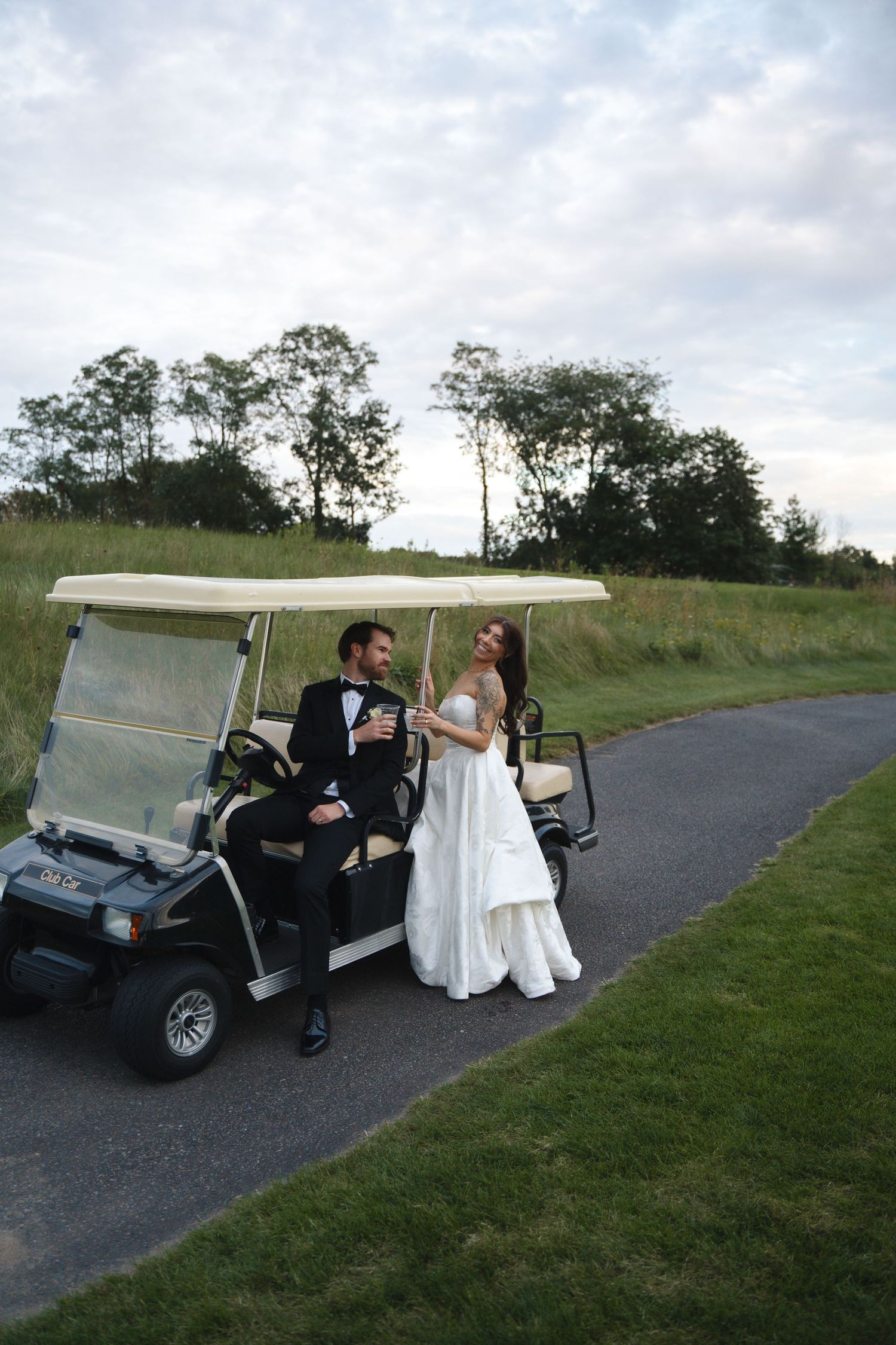 Bride and groom in wedding attire standing together beside a golf cart on a scenic outdoor path.