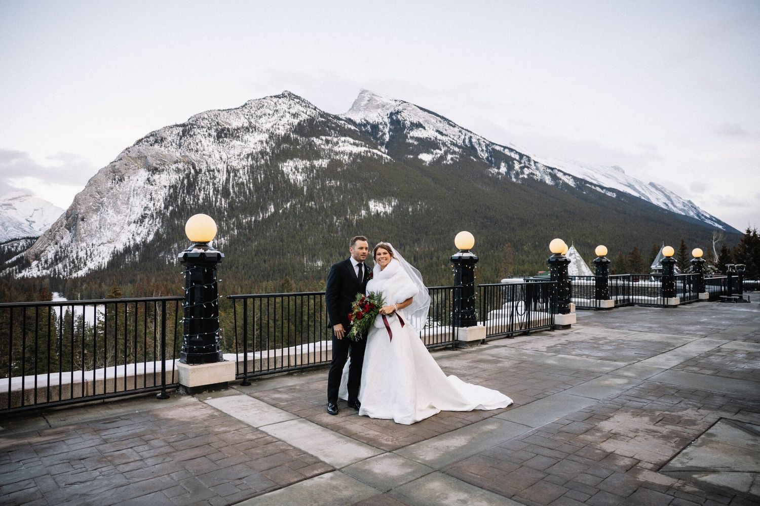 Bride in a white wrap and groom on the Fairmont Banff Springs terrace beneath a snow-dusted mountain in December