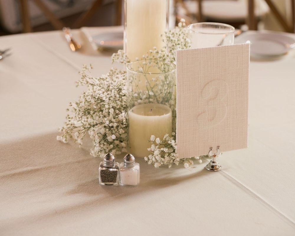 Elegant wedding table centerpiece with baby's breath, candles, and a linen table number card on white tablecloth.