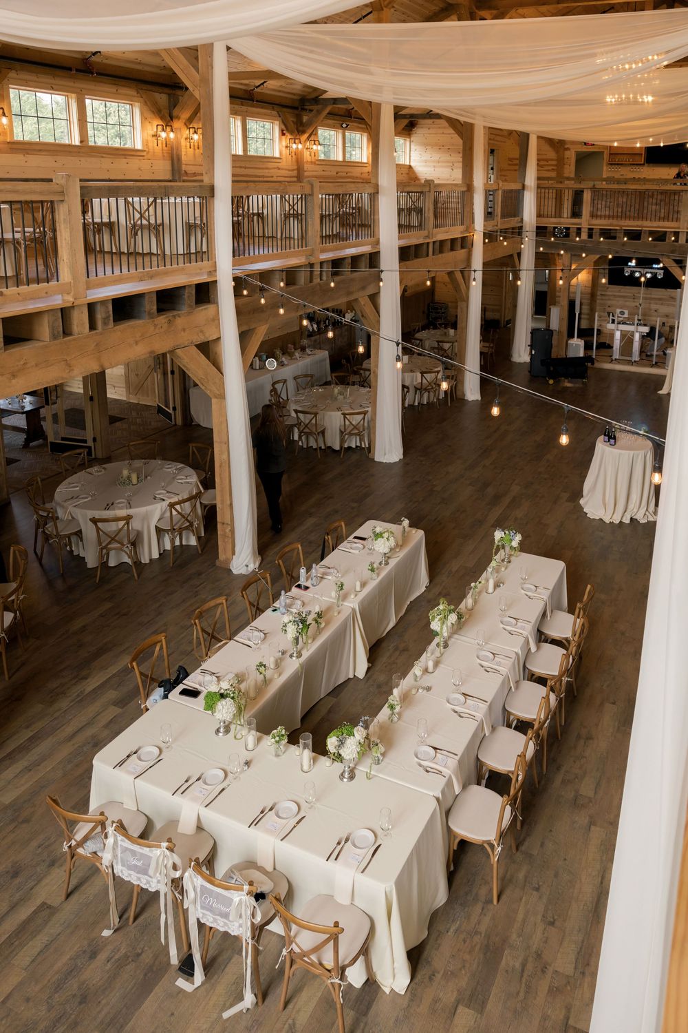 Elegant barn wedding reception with long white-draped tables, string lights, wooden columns, and a rustic two-story interior.