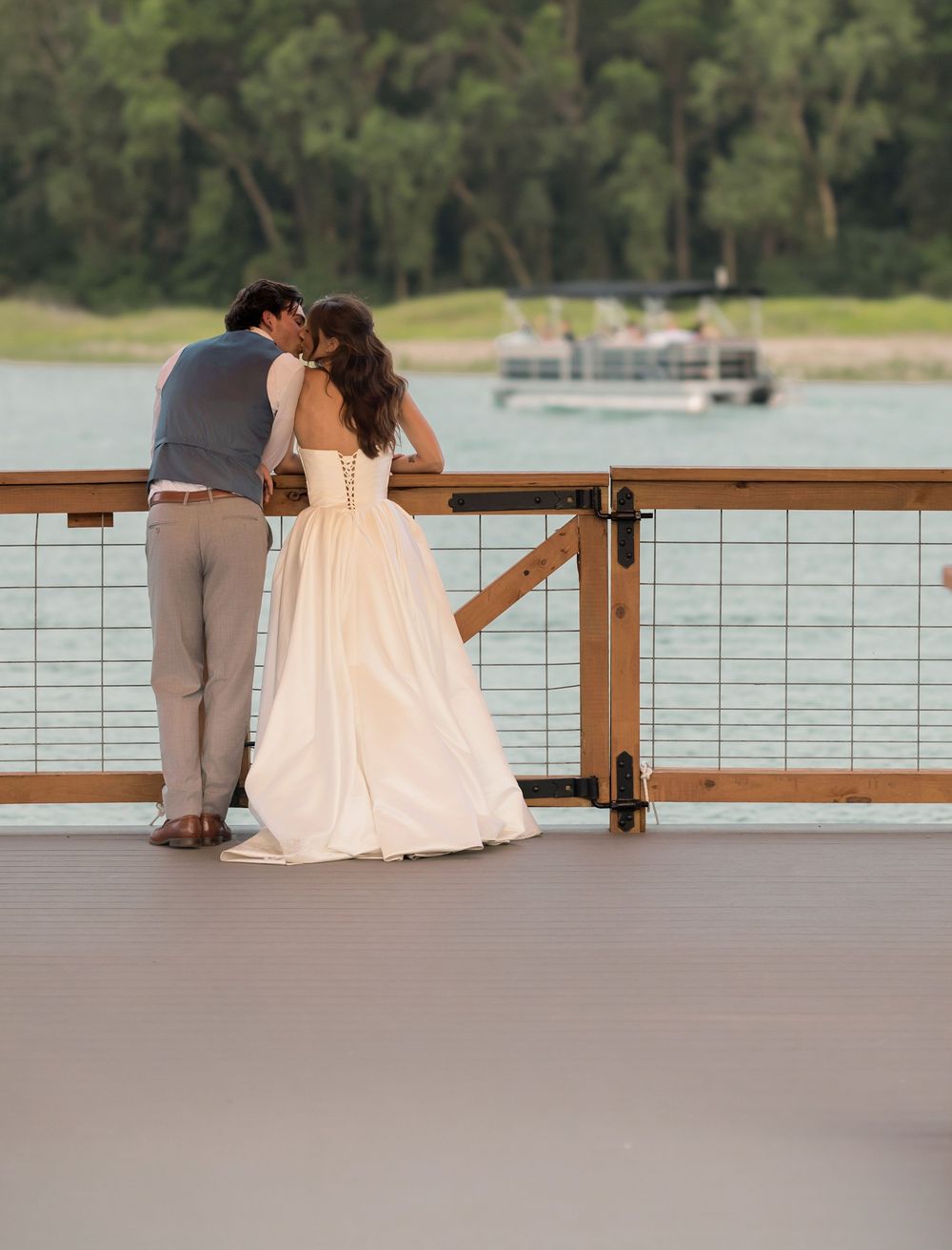 Bride and groom sharing a kiss on a waterfront dock, with a boat and lush green trees in the background.