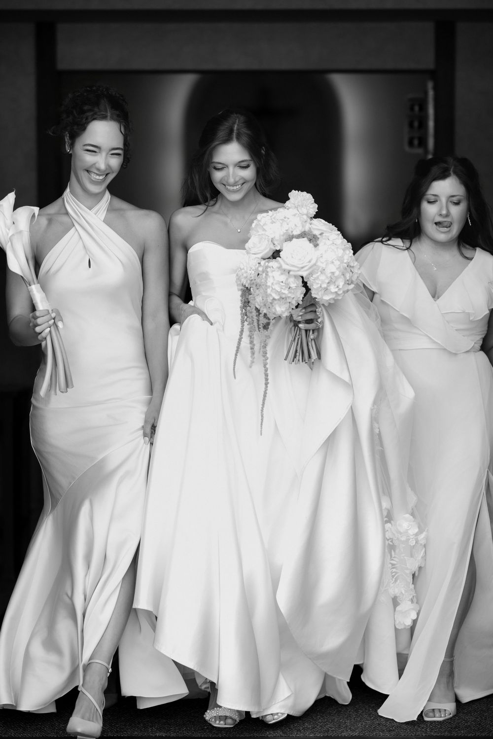 Black and white photo of a bride in a strapless gown holding white bouquet, flanked by two bridesmaids in elegant dresses.
