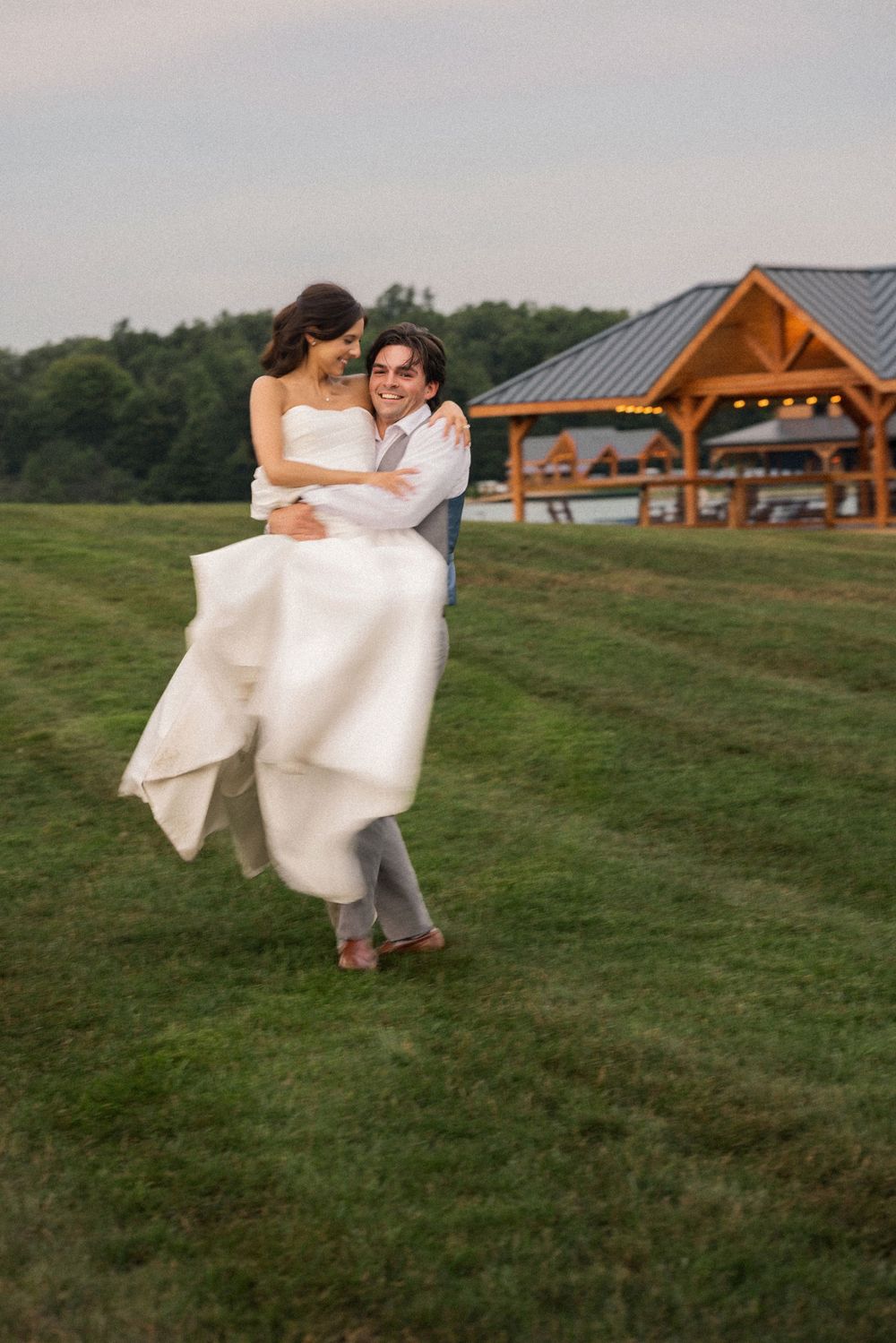 Groom carries smiling bride in white wedding dress across green lawn with rustic wooden pavilion in background.