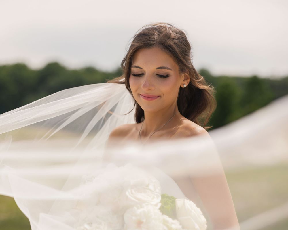 Elegant bride in white gown holding bouquet with veil flowing in wind, soft romantic outdoor wedding portrait.