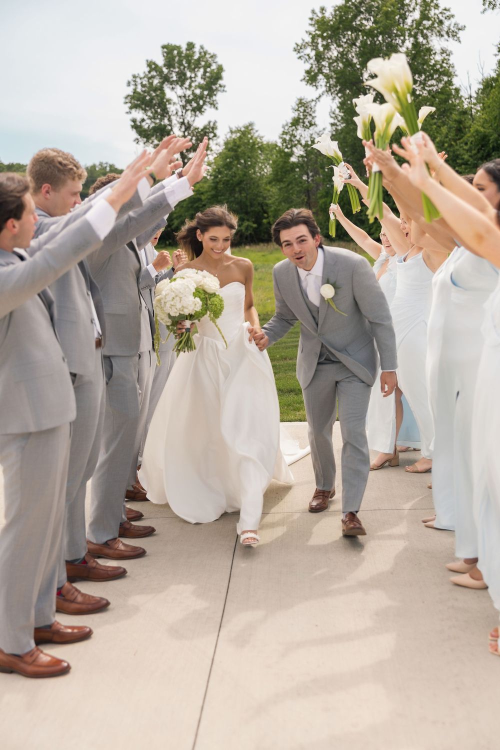 Bride and groom walk through arch of raised hands formed by wedding party in gray suits and light blue dresses outdoors.