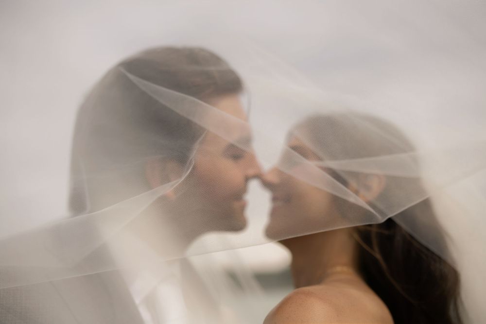 Bride and groom sharing a romantic moment under a flowing wedding veil, noses touching and smiling.