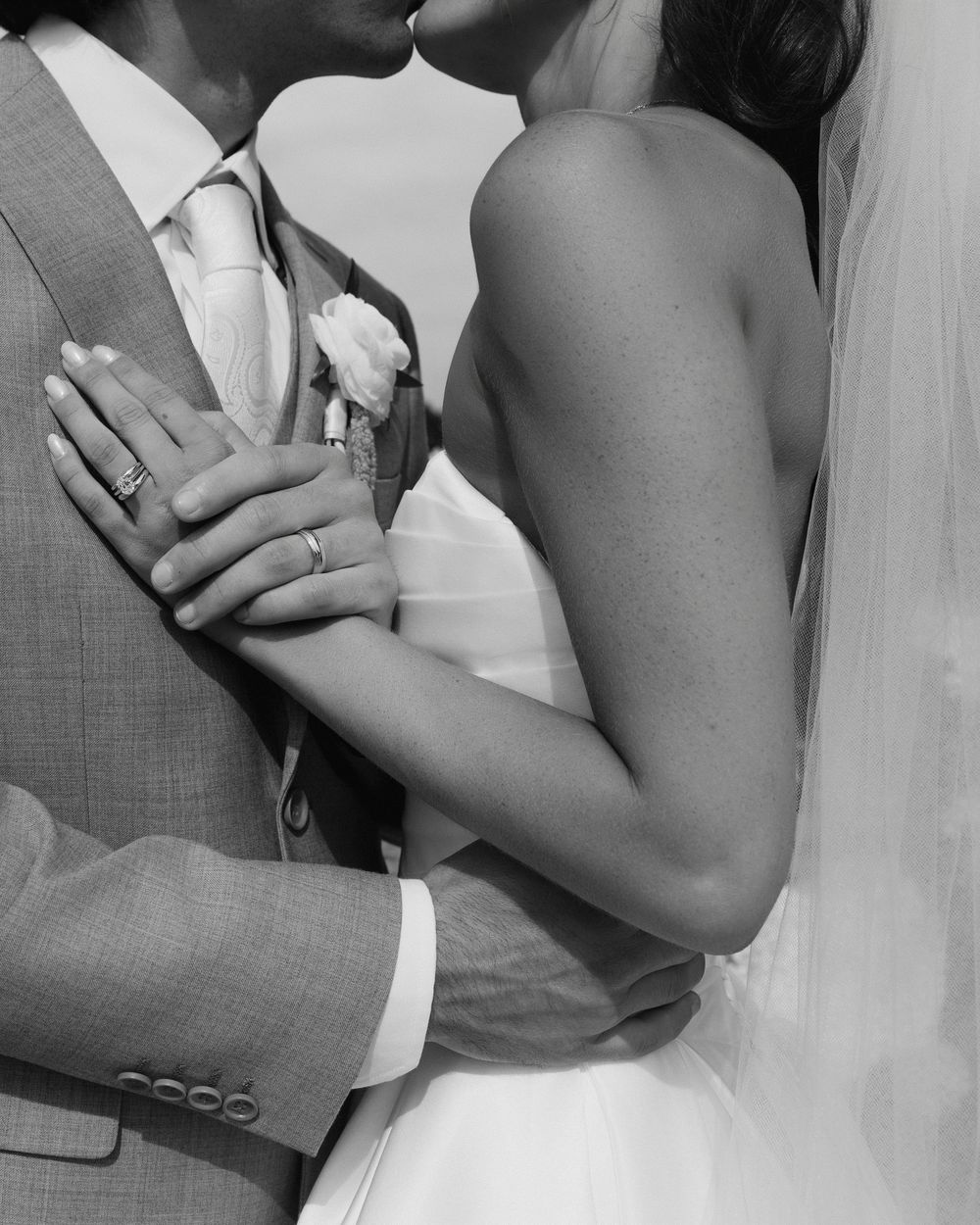 Black and white wedding photo of bride and groom kissing, showing rings and boutonniere.