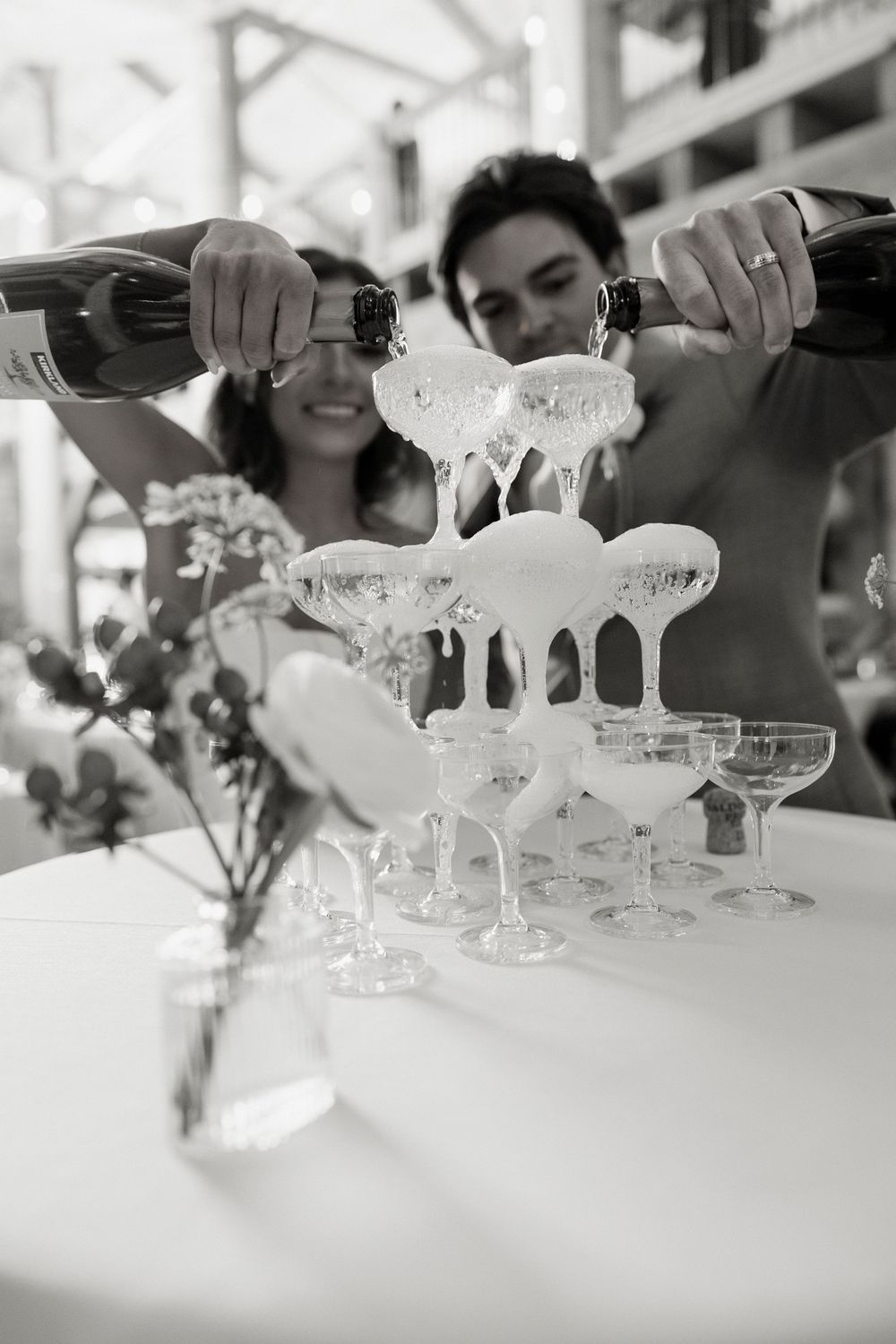 Couple pouring champagne into a tower of coupe glasses at a wedding reception in black and white.